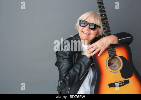 Ältere Frau in Lederjacke und Sonnenbrille Studio stehen isoliert auf Grau umarmen Gitarre träumen heiter Stockfoto