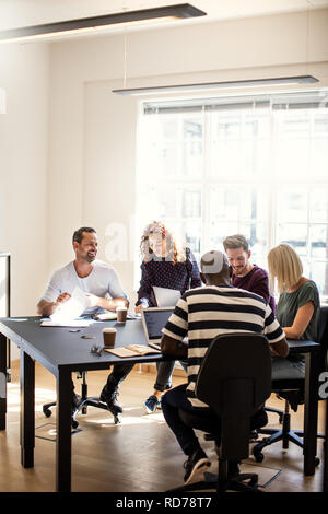 Vielfältige Gruppe von lächelnden jungen Designern zusammen, die um einen Konferenztisch in einem Büro Stockfoto
