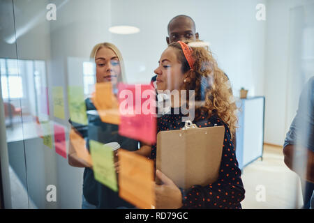 Junge Unternehmerin und ihr Team lesen Haftnotizen auf ein Glas Wand während eines Brainstormings in einem Büro Stockfoto