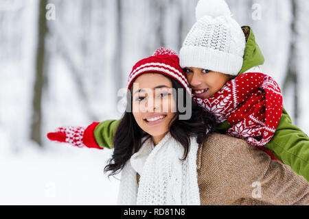 Attraktive afrikanische amerikanische Mutter huckepack fröhliche Tochter, lächelnd und mit Blick auf die Kamera im Winter Park Stockfoto