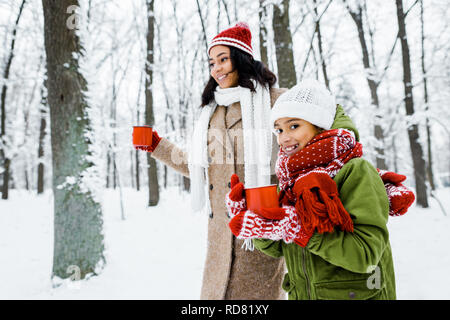 Attraktive afrikanischen amerikanischen Mutter und niedlichen Tochter zu Fuß und mit roten cups im verschneiten Wald Stockfoto