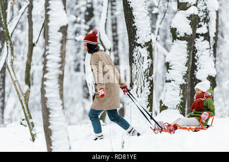 Afrikanische amerikanische Tochter mit der Mutter reiten auf Schlitten in der Nähe von Bäumen im verschneiten Wald Stockfoto