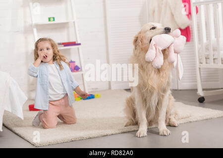 Adorable kid in der Nähe von Golden Retriever holding Spielzeug im Kinderzimmer sitzen überrascht Stockfoto