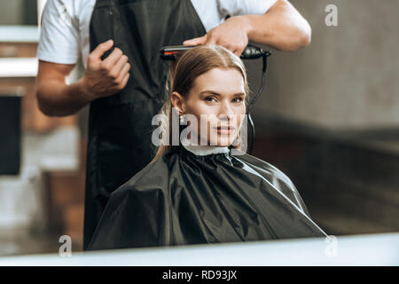 7/8 Schuß von Hairstylist mit Haarglätter und Mädchen an Spiegel im Beauty Salon suchen Stockfoto