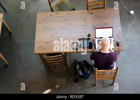 Ein Mann im t-shirt, der an einem Notebook arbeitet eng beobachten Veränderungen auf dem Laptop Anzeige bei rechteckigen Holztisch sitzen in einem lokalen beliebte Kaffee s Stockfoto