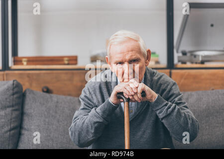 Verärgert pensionierter Mann mit grauen Haaren sitzen mit Spazierstock im Wohnzimmer Stockfoto