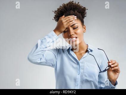 Müde Mädchen leiden unter schmerzhaften Kopfschmerzen und Stress. Foto der afrikanischen amerikanischen Frau in Blau Shirt die Hand auf die Stirn auf grauem Hintergrund. Medi Stockfoto