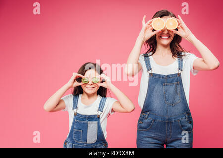 Glückliche Mutter und Tochter holding Früchte über Augen isoliert auf Rosa Stockfoto