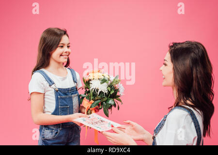 Fröhliche Tochter geben Karte und Blumen am Muttertag isoliert auf Rosa und Mom Stockfoto