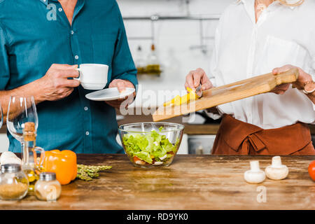 Zugeschnittenes Bild der reifen Frau, Gemüse in der Schüssel und Ehemann holding Tasse Tee in der Küche Stockfoto