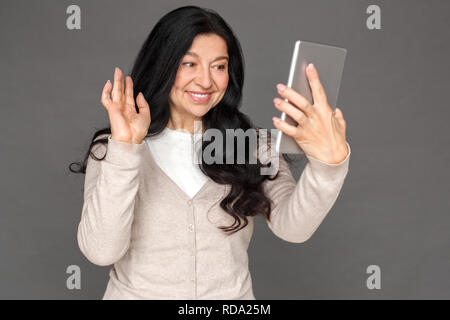 Freestyle. Reife Lady stehen isoliert auf Grau in Video Call winken in die Kamera freundlich Stockfoto