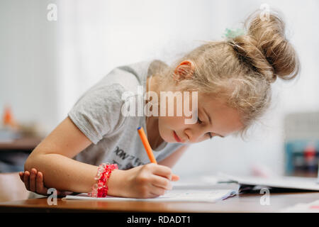 Ein siebenjähriges Mädchen sitzt zu Hause am Tisch und schreibt in einem Notebook, Abschluss einer Aufgabe lernen oder Wiederholen von Lektionen Stockfoto