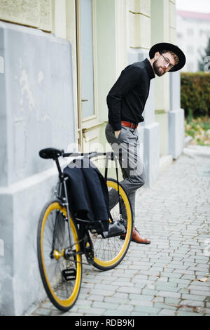 Volle Länge des selbstbewussten jungen bärtigen Mann Anpassung der Brille und Hut und Suchen an der Kamera, während in der Nähe von seinem Fahrrad im Freien stehend Stockfoto