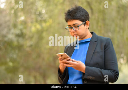 Closeup Portrait von selbstbewussten jungen indischen Unternehmen professionelle Frau mit kurzen Haaren, Kontrolle ihr Telefon in einem freien trägt einen schwarzen Anzug Stockfoto