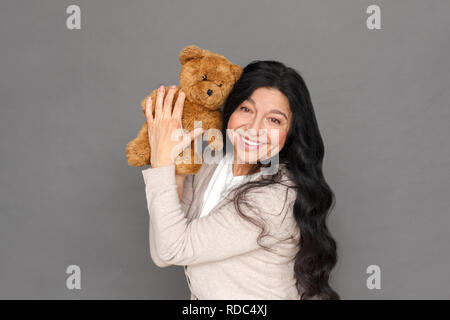 Freestyle. Reife Lady stehen isoliert auf Grau mit Teddybär bis glücklich Stockfoto