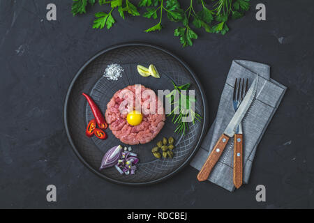 Steak Tartar mit Eigelb und Zutaten aus schwarzer Keramik Teller, Besteck Messer und Gabel auf schwarzen Stein Beton texturierte Oberfläche Hintergrund. Cop Stockfoto