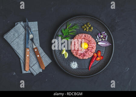 Steak Tartar mit Eigelb und Zutaten aus schwarzer Keramik Teller, Besteck Messer und Gabel auf schwarzen Stein Beton texturierte Oberfläche Hintergrund. Cop Stockfoto