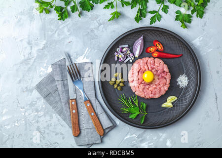 Steak Tartar mit Eigelb und Zutaten aus schwarzer Keramik Teller, Besteck Messer und Gabel auf hellen grauen Stein Beton texturierte Oberfläche Hintergrund Stockfoto
