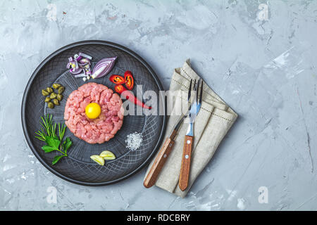 Steak Tartar mit Eigelb und Zutaten aus schwarzer Keramik Teller, Besteck Messer und Gabel auf hellen grauen Stein Beton texturierte Oberfläche Hintergrund Stockfoto