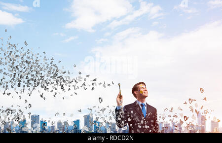 Porträt der jungen nachdenklicher Mann mit Pinsel in der Hand und Wegsehen im Stehen mit fliegenden Buchstaben und gegen moderne Stadtbild anzeigen Stockfoto