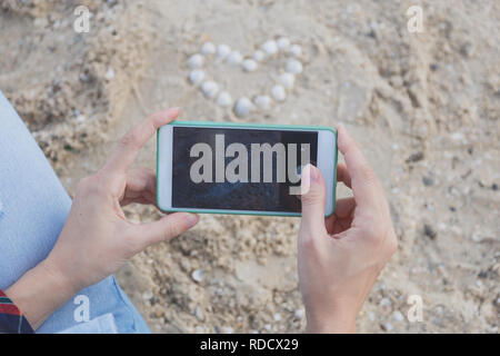 Weibliche Hände mit Smartphones Bilder von Muscheln auf dem Sand in der Form eines Herzens. Stockfoto
