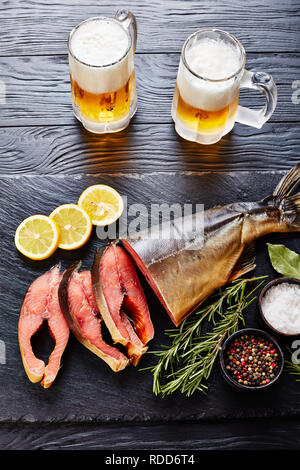 Wildlachs schnitt in Steaks mit frischem Rosmarin Salz und Pfeffer auf einem schwarzen Stein Fach mit frisch geschäumter Bier im Glas Tassen, vertikale Ansicht von oben Stockfoto