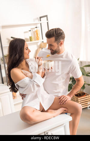 Lächelnde Frau füttern Mann mit Müsli beim Frühstück am Morgen Stockfoto