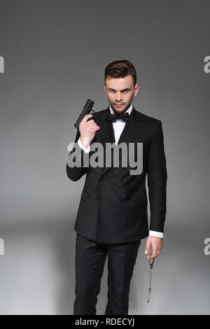 Stattliche Secret Agent im Tuxedo holding Pistole und Handschellen, isoliert auf Grau Stockfoto