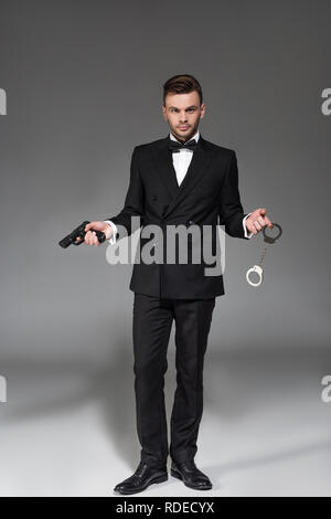 Elegante Secret Agent im Tuxedo holding Pistole und Handschellen auf Grau Stockfoto