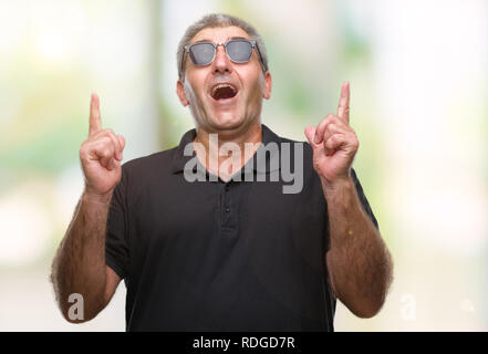 Gut aussehender älterer Mann Sonnenbrille tragen über isolierte Hintergrund erstaunt und überrascht mit der Suche und zeigt mit Fingern und erhobenen Armen. Stockfoto
