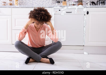 Sorgen junge Frau sitzt Vor der beschädigten Geschirrspüler mit Schaum Aus Aus Stockfoto