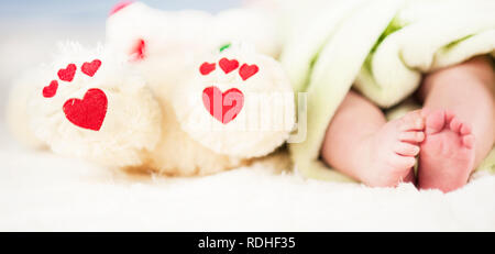 Kleine zarte Beine des Babys, zusammen mit den Pfoten eines Teddybär mit Herz auf einer weißen Decke Stockfoto