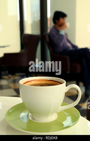 Eine Tasse heißen Kaffee auf einem weißen Tisch mit verschwommenen Kaffee trinken Mann im Hintergrund Stockfoto