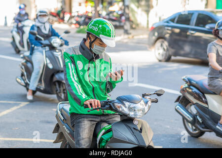 Danang, Vietnam - Oktober 14, 2018: Ein junger Mann auf einen Roller, trägt einen grünen Helm und Jacke, schaut auf sein Handy. Stockfoto