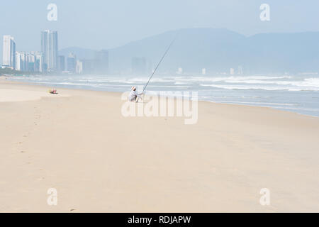 Danang, Vietnam - November 1, 2018: ein Mann Fische mit einem sich drehenden Stange in den flachen wellige Meer sitzen auf dem Sonnenbeschienenen leeren Strand mit Danang City hinter Stockfoto