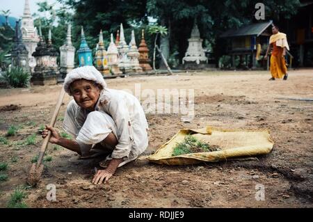 Attapu/Laos - JUL 06 2011: ältere Frau Aufräumen der Hinterhof eines Klosters, während ein Mönch ist vorbei im Hintergrund Vor vielen kleinen Stockfoto