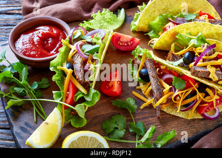 Tacos mit gebratenem Rindfleisch in Streifen schneiden, Salatblätter, roten Zwiebeln, Tomaten, Oliven und shredded Cheddar Käse auf einem Schneidebrett mit Tomate sals Stockfoto