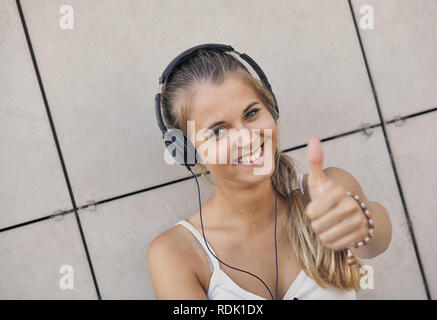 Junge lächelnde Frau in einem weißen T-Shirt mit Musik hören und zeigt den Daumen nach oben Stockfoto