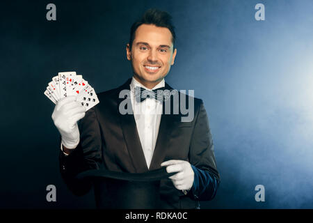 Berufliche Tätigkeit. Magier in Anzug und Handschuhe isoliert stehend an der Wand zeigen Trick mit Karten und Hut lächelnd glücklich Stockfoto