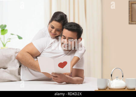 Glückliches junges Paar, die den Valentinstag feierten in einem Schlafzimmer, umarmen und gemeinsam lesen Grußkarte mit einem großen Herzen Form Stockfoto