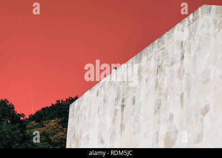 Ein Mann sitzt auf der Kante einer Mauer aus Stein mit dem Coral farbigen Himmel im Hintergrund und einigen Bäumen Stockfoto