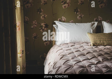 Innenansicht Schlafzimmer mit Vorhängen und Tapeten mit Blumenmuster, pale pink Quilt und weißen Kopfkissen auf dem Doppelbett. Stockfoto