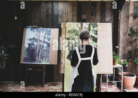 Ältere Frau mit schwarzen und weißen Schürze stehen im Studio, arbeiten auf der Malerei der Bäume im Wald. Stockfoto