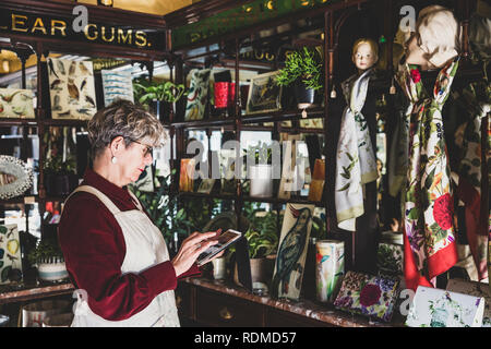 Ältere Frau Brille, rotes Kleid und der weißen Schürze in Interior Design Store, digitale Tabelle. Stockfoto