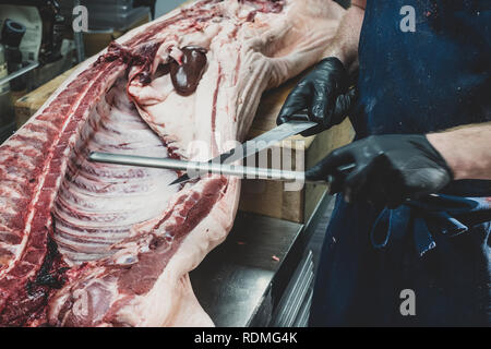 Nahaufnahme von Metzger mit schwarzen Gummi Handschuhe an einem geschlachtete Schweine auf der Butcher Block, Schärfen der Messer. Stockfoto