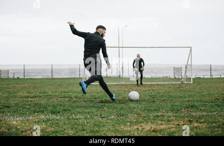 Der Fußballer schlägt einen Elfmeterschuss. Fußballspieler tritt den Ball in Richtung Torpfosten, während sich der Torwart in Position befindet. Stockfoto