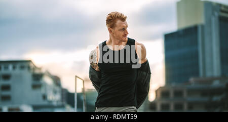 Fitness-Mann macht sich bereit für das Training und zieht sein Sweatshirt aus. Mann mit muskulösem Körper, der auf dem Dach steht und sein Hemd für Fitnesstraining auszieht. Stockfoto