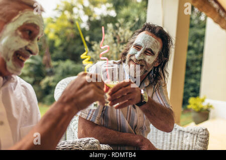 Gerne alte Menschen toasten Saft Gläser mit Gesichts Clay Mask auf. Lustig ältere Männer sitzen zusammen mit Gesichtsmaske mit Saft. Stockfoto