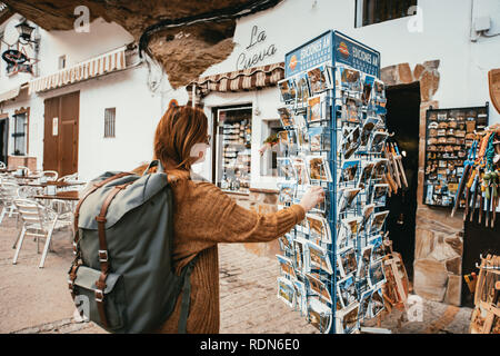 Junge Frau mit einem grünen Rucksack, orange Jacke und rote Haare blickt auf eine Postkarte stand eines Kiosk in einem kleinen spanischen Dorf bauen in einem Rock. Stockfoto