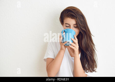 Junge brünette Frau im weißen T-Shirt trinken aus blau Tasse mit beiden Händen an der Kamera, die mit einem freundlichen Lächeln. Stehend gegen weiße Wand wit Stockfoto